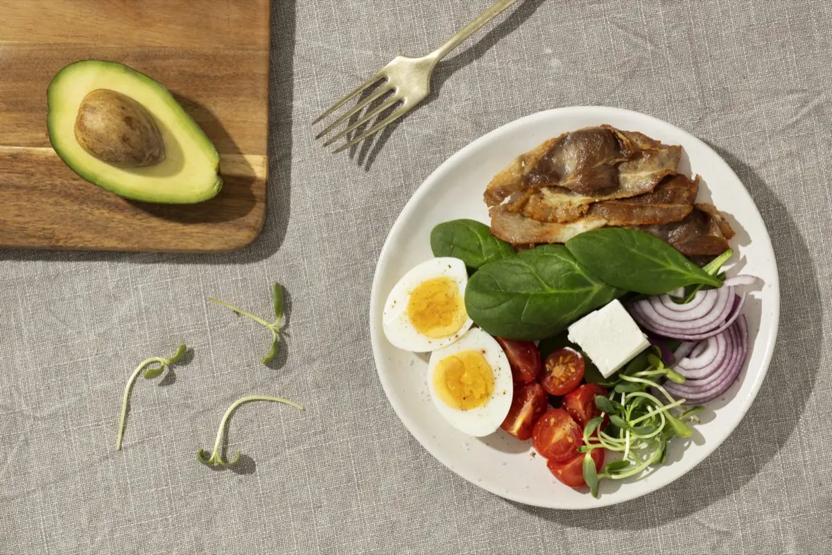 Plate with eggs, bacon, spinach, tomato, feta, and onion beside an avocado